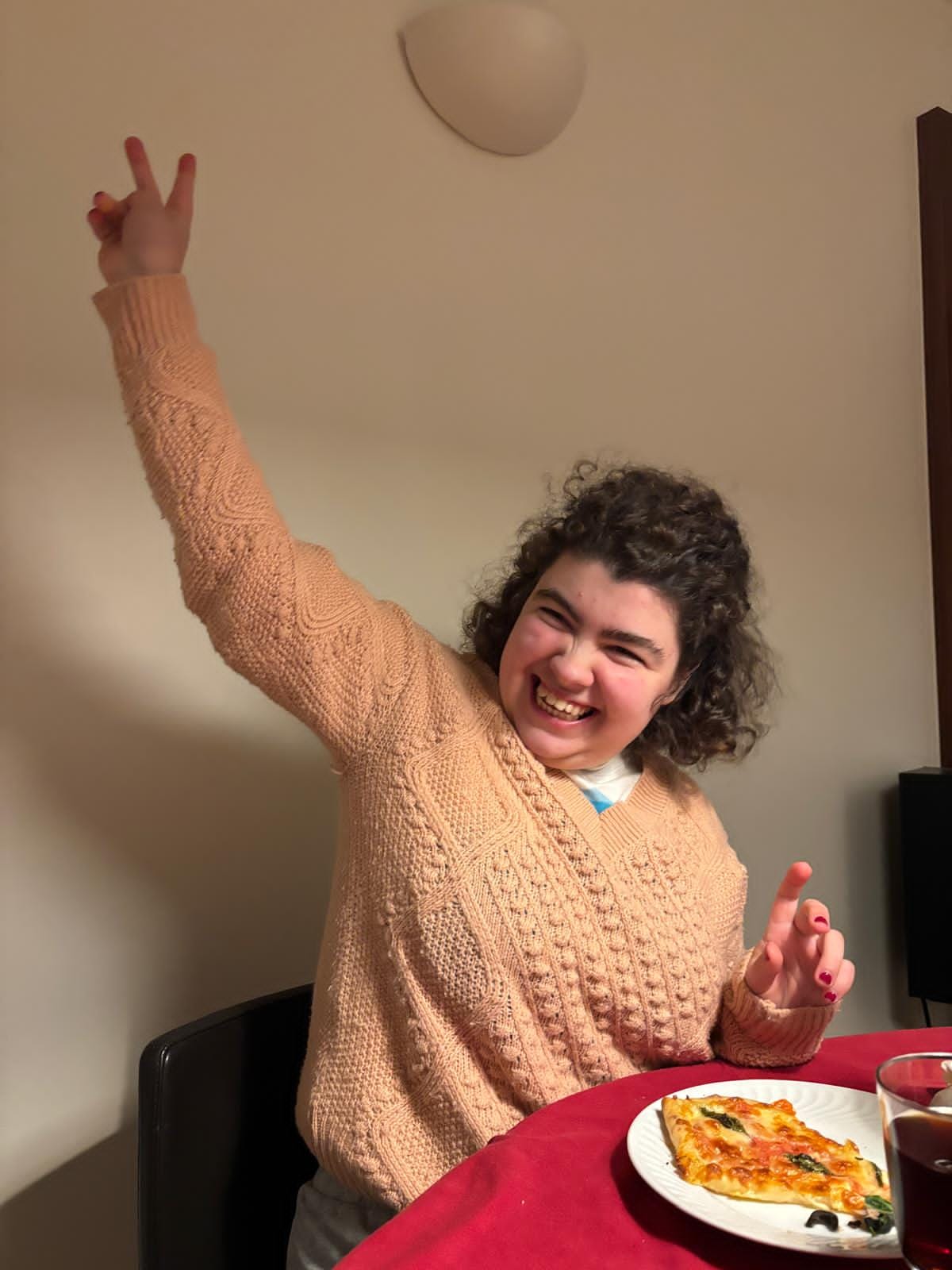 Leonor, a young adult, at a table with a slice of pizza, making a peace sign